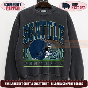 May include: A charcoal gray crewneck sweatshirt featuring a graphic print with the text SEATTLE 1976 FOOTBALL. The design includes a dark blue football helmet with a face mask, outlined in light green, centered on the chest of the garment.