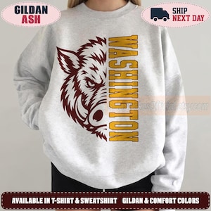 May include: A light grey crewneck sweatshirt featuring a maroon graphic of a boar head on the left side and the word WASHINGTON printed vertically in bold yellow block letters on the right side of the chest area.