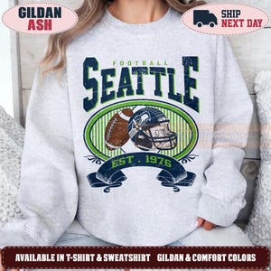 May include: A light gray crewneck sweatshirt featuring a distressed graphic print. The design includes the text SEATTLE FOOTBALL EST. 1976, a brown football, and a dark blue and green football helmet centered within a circular green and blue frame.