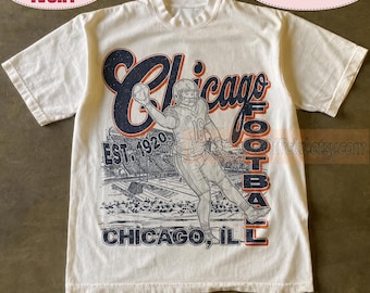 May include: An ivory short-sleeve t-shirt featuring a graphic print of a football player throwing a ball. The design includes the text Chicago, Football, Est. 1920, and Chicago, IL in navy blue and orange lettering over a stadium background.