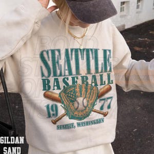 May include: A Gildan Sand sweatshirt featuring a teal and brown graphic design. The design includes the words "SEATTLE BASEBALL 1977" with a baseball glove, baseball, and crossed bats. The text "SEATTLE, WASHINGTON" is also included.