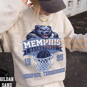 May include: A sand-colored sweatshirt featuring a blue and white graphic. The graphic includes a snarling bear, the words "Memphis Basketball," a basketball going through a net, and the numbers "19" and "95." The text "Memphis, Tennessee" is also displayed.