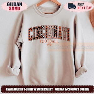 May include: A sand-colored crewneck sweatshirt featuring the word CINCINNATI in large, block-style letters with varied patterns including checkers, stripes, and leopard print. Below, the word FOOTBALL and a small football graphic are printed.