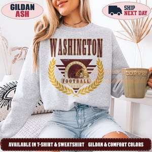 May include: A light gray crewneck sweatshirt featuring a maroon and gold graphic on the chest. The design includes the text WASHINGTON above a triangular emblem with a football helmet, the word FOOTBALL, the year 1932, and a gold laurel wreath.