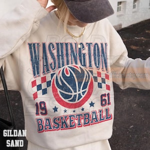 May include: Cream-colored sweatshirt with a distressed graphic featuring "WASHINGTON BASKETBALL 19 61." The design includes a basketball, stars, and checkered flags in red and blue. The sweatshirt is labeled "GILDAN SAND."