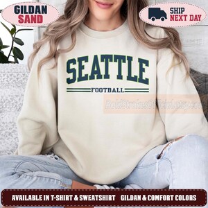 May include: A sand-colored crewneck sweatshirt featuring the word SEATTLE in large, dark navy blue collegiate-style lettering. Below the text, the word FOOTBALL is printed in a smaller font, flanked by two horizontal dark navy blue lines.
