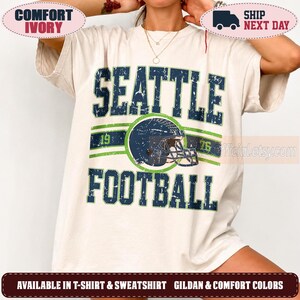 May include: An ivory short-sleeve t-shirt featuring a distressed navy blue and green graphic. The design includes the text SEATTLE FOOTBALL, the numbers 19 and 76, and a central illustration of a football helmet with a face mask.