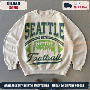 May include: A sand-colored crewneck sweatshirt featuring a graphic print with the text SEATTLE 1976 and Football. The design includes a large green football illustration containing a white silhouette of the Seattle skyline and the Space Needle.