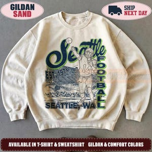 May include: A sand-colored crewneck sweatshirt featuring a graphic print of a football player in a helmet and jersey with the number 14. The design includes the text Seattle Football, Est. 1976, and Seattle, WA, over a stadium background.