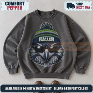May include: A charcoal gray crewneck sweatshirt featuring a graphic of an eagle wearing black sunglasses and a blue, green, and gray knit beanie with a pom-pom. The beanie has a patch with the text SEATTLE centered on the front of the garment.