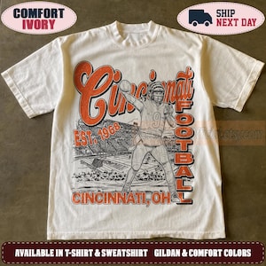 May include: An ivory short-sleeve t-shirt featuring a graphic print of a football player in a stadium. The text reads Cincinnati Football, Est. 1968, and Cincinnati, OH. The design uses orange and black ink with a distressed, vintage-style aesthetic.