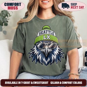 May include: A moss green short-sleeve t-shirt featuring a graphic of a bird wearing a lime green and white beanie with the text SEATTLE LX and dark sunglasses. The bird has blue and white feathers and a sharp beak, centered on the chest.