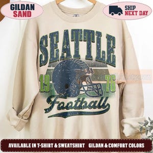 May include: A sand-coloured crewneck sweatshirt featuring a distressed graphic print. The design includes the text SEATTLE in dark blue, the number 1976 in green, and the word Football in dark blue, centred around a dark blue football helmet.