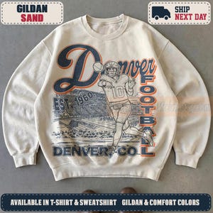 Puede incluir: Una sudadera de cuello redondo color arena con un estampado gráfico de un jugador de fútbol americano con casco y camiseta número 10, un balón, un estadio y el texto Denver Football Est. 1960 en tinta azul marino y naranja en el frente.