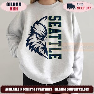 May include: A light gray crewneck sweatshirt featuring a dark blue graphic on the front. The design consists of a stylized eagle head on the left side and the word SEATTLE printed vertically in a bold, block font on the right side.