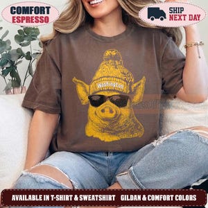 May include: A brown short-sleeved t-shirt featuring a yellow graphic of a pig wearing a knit beanie with the text Washington and black sunglasses. The shirt has a crew neckline and a relaxed fit, showcasing a detailed illustration of the pig.