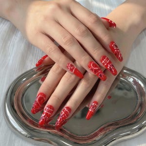 Pode incluir: Unhas artificiais longas, vermelhas, em forma de caixão com um desenho paisley branco. As unhas estão numa bandeja prateada. As unhas são de cor vermelha vibrante com um padrão branco.