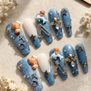 Puede incluir: Un conjunto de uñas postizas largas con un diseño similar al denim. Cada uña está adornada con varios abalorios, incluyendo osos de peluche, delfines, estrellas, cruces y detalles plateados. Algunas uñas tienen una base blanca.
