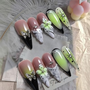 May include: A collection of long, almond-shaped artificial nails with various designs. Some nails feature butterfly accents, floral embellishments, and rhinestone details. The color palette includes pink, black, white, and green, with gradient effects.