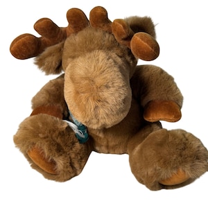 Può includere: Un peluche a forma di alce con pelliccia marrone, corna e zoccoli. L'alce ha una sciarpa verde e bianca intorno al collo. Il giocattolo è seduto e ha un'espressione amichevole.