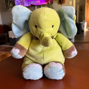 May include: A plush elephant toy in shades of yellow, blue, and brown. The elephant has large blue ears, brown accents on its feet and arms, and small black eyes. The toy is sitting on a brown surface.