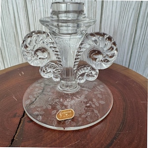 Puede incluir: Candelabro de cristal transparente con un diseño floral grabado en la base. El candelabro presenta una base acampanada, un tallo central y adornos en espiral. Una pequeña etiqueta con la palabra "Fostoria" es visible.
