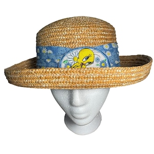 May include: A straw hat with a wide brim and a blue band featuring a daisy pattern. The hat has a circular patch with Tweety Bird and a daisy. The hat is displayed on a white mannequin head.