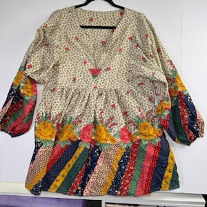 May include: A long-sleeved tunic featuring a cream-colored bodice with a small red floral print, a V-neckline, and a wide band of large yellow and pink roses above a hemline composed of diagonal, multi-colored striped fabric patches.