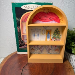 Puede incluir: Una estantería decorativa amarilla de dos niveles con un diseño de escenario teatral. La parte superior presenta una cortina roja con el texto "The Nutcracker Ballet". Los estantes muestran una escena de escenario con un árbol de Navidad y una escena de ballet.