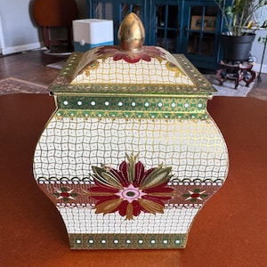 May include: A decorative, vintage metal container with a hinged lid. The container is white with a gold, green, and burgundy floral design. The lid has a gold finial. The container is approximately 7 inches tall and 6 inches wide.