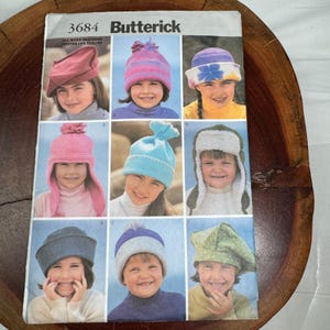May include: A Butterick 3684 sewing pattern envelope featuring illustrations of nine different hat styles. The designs include berets, beanies with pom-poms, earflap hats with faux fur lining, and brimmed hats in various colors like pink, blue, and green.