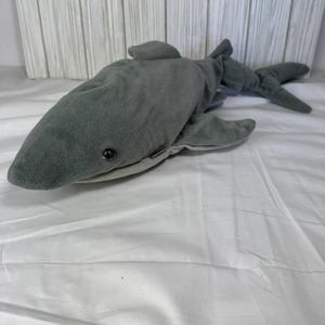 May include: A plush shark toy featuring a soft, dark gray exterior with a lighter gray underbelly. The stuffed animal has a pointed snout, a single visible black plastic eye, prominent side fins, and a detailed tail fin structure.