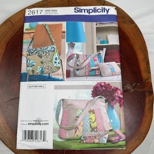 Puede incluir: Un sobre de patrón de costura Simplicity 2617 con ilustraciones de tres diseños diferentes de bolsos de hombro con detalles de tela de patchwork, patrones florales y pequeños bolsos a juego. El nombre de la marca y texto son visibles.
