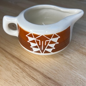 May include: A small cream pitcher made of off-white ceramic featuring a wide horizontal band of burnt orange glaze. The center of the orange band displays a white geometric pattern consisting of stacked triangles and vertical lines.