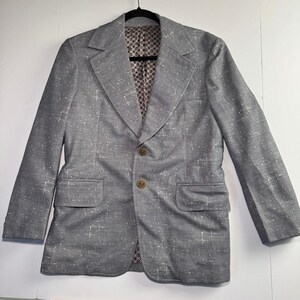 May include: A grey blazer featuring a white cross-hatch pattern, wide lapels, and two gold-toned buttons on the front. The interior lining displays a repeating brown and white geometric pattern. The jacket includes two front flap pockets.