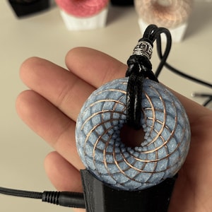 May include: A blue and copper-colored pendant with a spiral design, held in a hand. The pendant is attached to a black cord and sits on a black base. Other similar pendants in various colors are visible in the background.