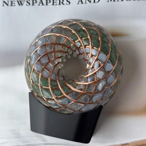 May include: A decorative sphere with a copper wire grid, filled with blue and green stones, sits on a black stand. The sphere has a hole in the center. The background includes text from a newspaper that reads "NEWSPAPERS AND MAGAZ".