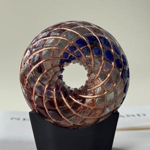 May include: A decorative, spherical object with a hole in the center, composed of a mosaic of purple, blue, and clear glass pieces, encased in a copper wire grid. It sits on a black, curved base.