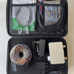 May include: Open black carrying case containing electronic components. Includes a silver amplifier, power adapters, cables, and a decorative sphere with copper wire and blue and brown accents. Also includes a remote control and other accessories.