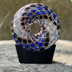 Pode incluir: Uma escultura toroidal feita de resina transparente incrustada com pedras de lápis-lazúli azul, peças de obsidiana preta e uma bobina de fio de cobre em espiral. O objeto circular é suportado por um suporte preto sólido e geométrico.