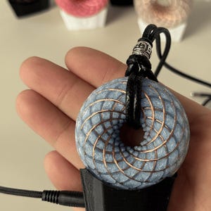 May include: A light blue, torus-shaped object with a copper wire coil pattern, suspended by a black cord with a silver-toned metal bead. The object is mounted on a black, rectangular base connected to a thin black power cable.