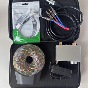 Könnte beinhalten: Ein schwarzes Etui mit Reißverschluss, das Audioausrüstung enthält. Enthält ein silbernes und schwarzes Kabel, ein silbernes und schwarzes Adapterkabel, eine silberne Metallbox, ein schwarzes Netzteil und eine dekorative Kugel mit Kupfer- und grünen Akzenten.