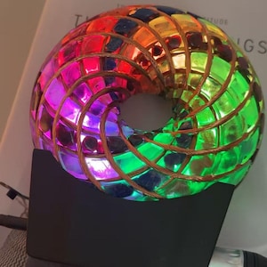 May include: A colorful, illuminated art piece shaped like a torus. The mosaic design features a spectrum of colors, including red, green, and purple. The piece is mounted on a black base and has a copper wire frame.