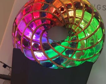 以下が含まれることがあります： トーラス型のカラフルな照明アート作品。モザイクデザインは、赤、緑、紫など、さまざまな色を特徴としています。黒いベースに取り付けられ、銅線フレームが付いています。