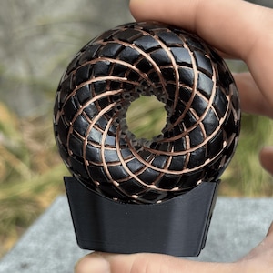Op de afbeelding: Een zwart en koperkleurige decoratieve bol met een geometrisch patroon. De bol wordt op zijn plaats gehouden door een zwarte basis. De bol heeft een holle kern en is gemaakt van onderling verbonden vormen.