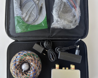 Könnte beinhalten: Ein schwarzer Transportkoffer mit Reißverschluss enthält eine kugelförmige Mosaiklampe mit blauen und grünen Glasfliesen, ein rechteckiges silbernes Elektronikgerät mit Goldanschlüssen, zwei schwarze Netzteile mit Kabeln und diverse Kabel.