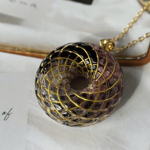 May include: A circular pendant featuring a clear resin torus shape filled with small, dark purple and black stone fragments. A gold-colored wire is wrapped in a spiral pattern around the exterior of the resin, attached to a gold-tone chain.