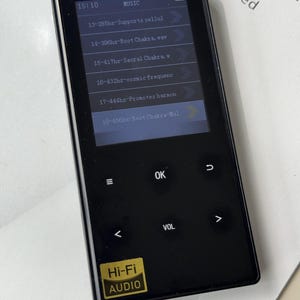May include: Black Hi-Fi audio player with a screen displaying a music menu. The screen shows options like "Root Chakra" and "cosmic frequenc". The player has control buttons and a "Hi-Fi AUDIO" sticker.
