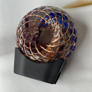 May include: A decorative, donut-shaped object with a copper wire cage filled with blue and purple stones. The object sits on a black stand, and the background is a white surface with the word "NEWS" visible.
