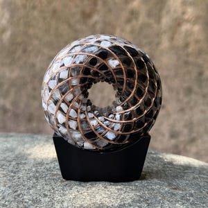 May include: A decorative, spherical object with a central hole, composed of black, white, and gray stones, encased in copper wire. It sits on a black, rectangular base, creating a unique, artistic display.